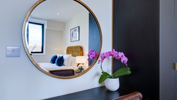 A hotel room with a round mirror, a bed with blue pillows, a nightstand with a lamp, and a vase with purple flowers. - Hybrid Resi - Cheam  (Sutton)