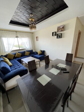 A modern living room with a dark wooden ceiling, a blue sectional sofa with yellow cushions, a glass dining table with chairs, and a large window with sheer curtains.