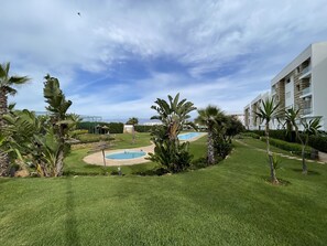 A well-maintained pool area with a clear sky, surrounded by lush greenery and modern buildings.