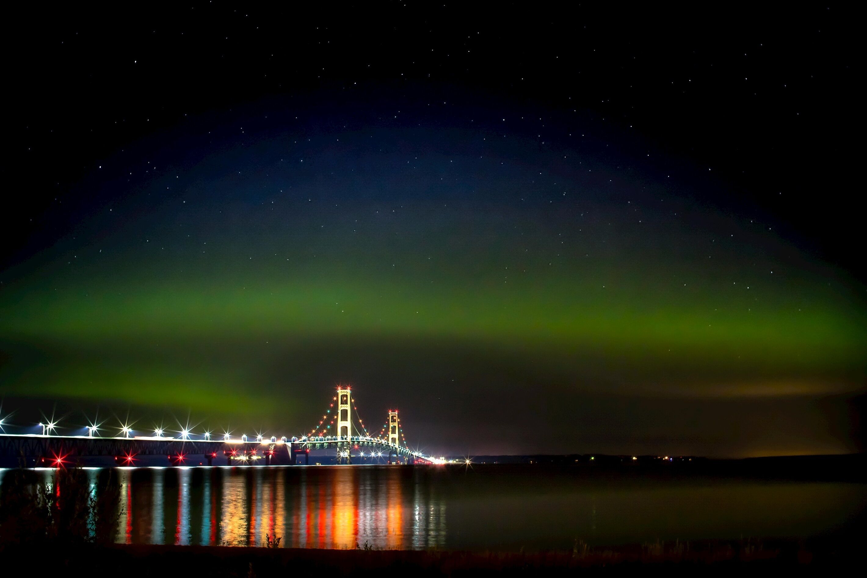 A bridge with lights reflecting on water, under a night sky with visible stars and green aurora.