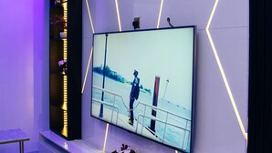 Uma sala de estar moderna com uma grande televisão de tela plana fixada na parede, uma estante embutida com objetos decorativos e um design minimalista com detalhes de iluminação geométrica.