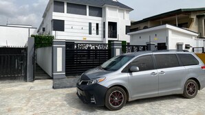 Uma casa moderna de dois andares com entrada fechada por portão, um carro estacionado em frente e céu claro.