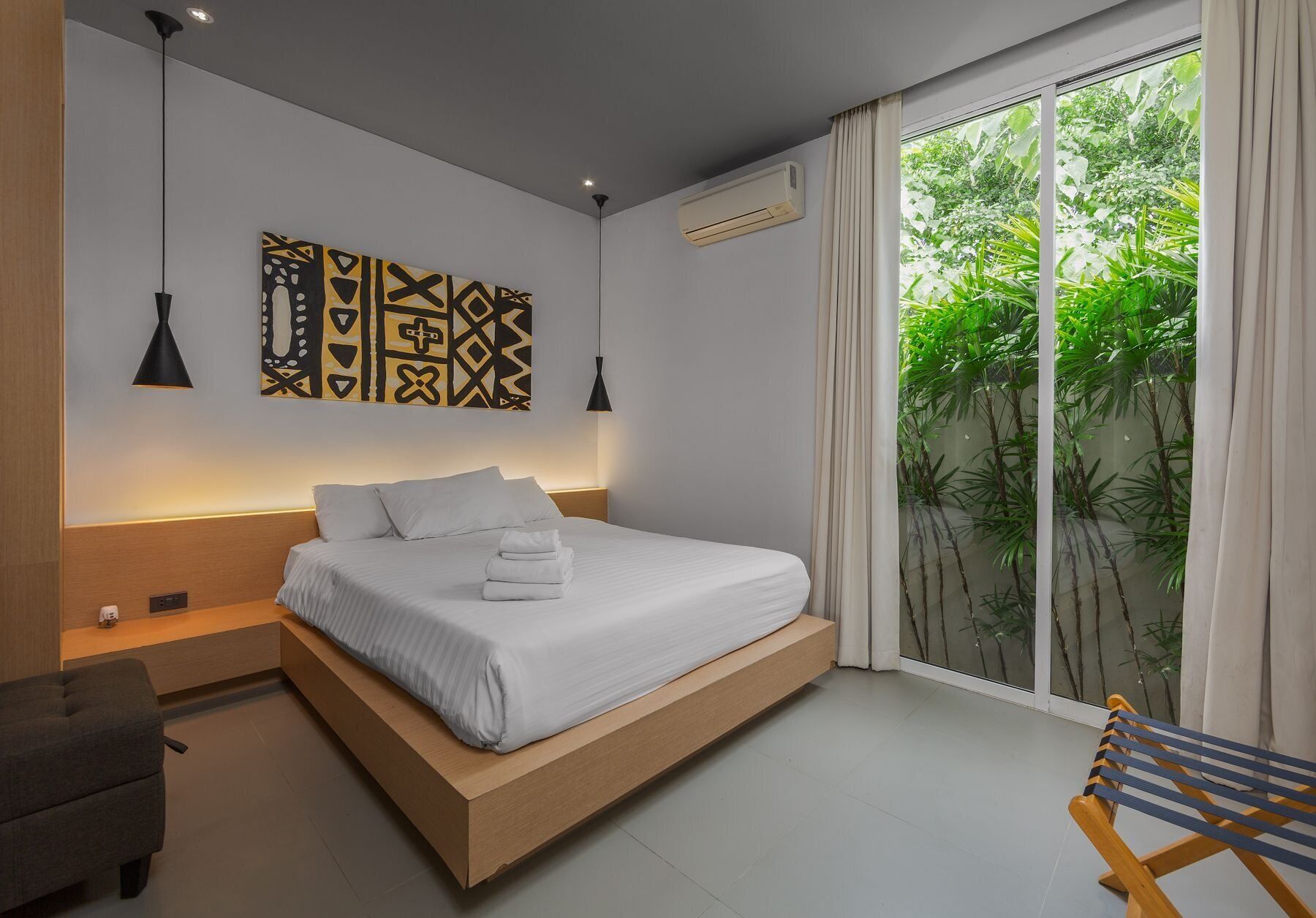 A modern bedroom with a bed, bedside tables, a bench, and a view of greenery through a sliding door.