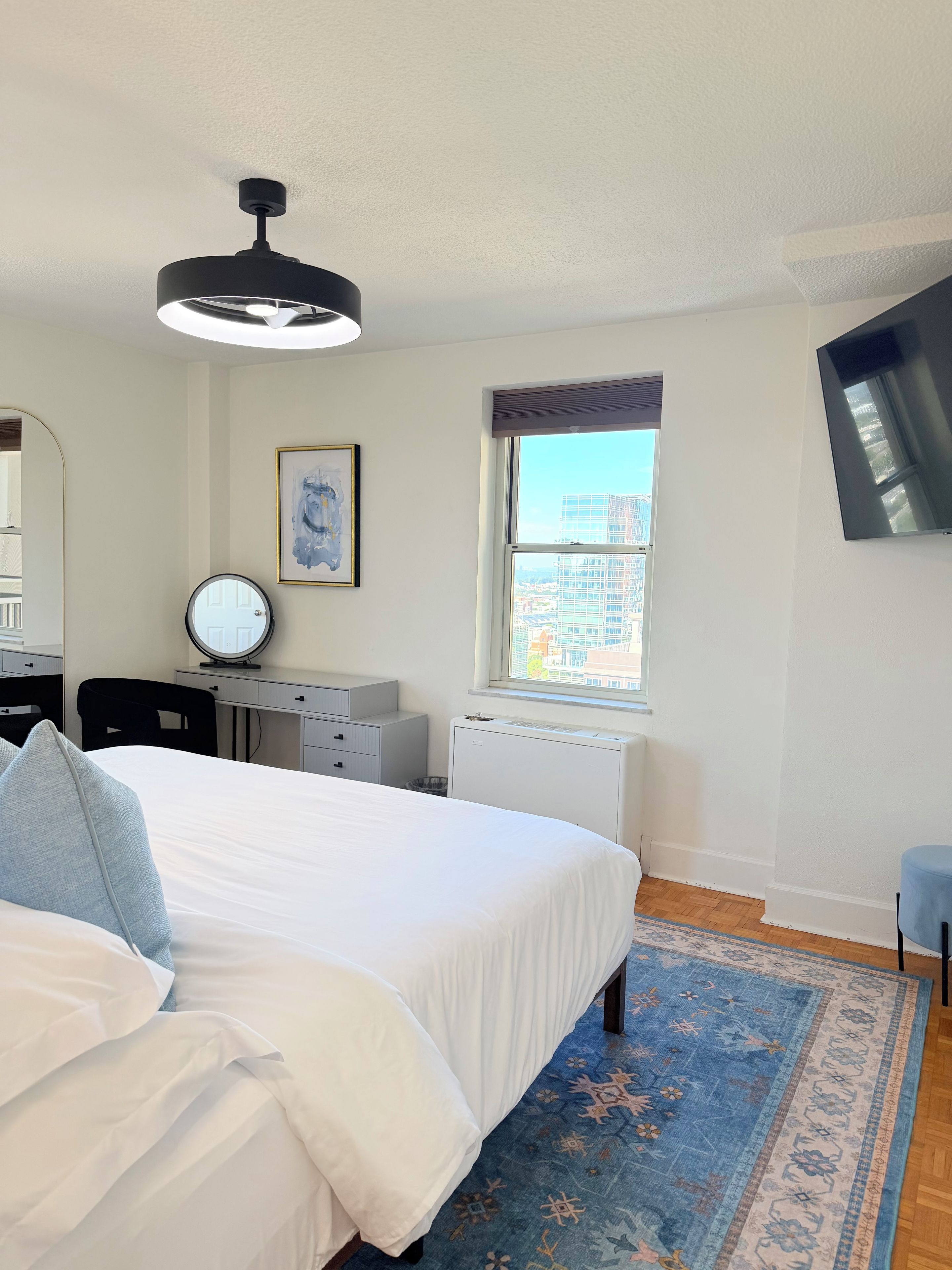 A bedroom with a bed, a desk, a chair, a radiator, a window with a view of buildings, and a mounted TV.