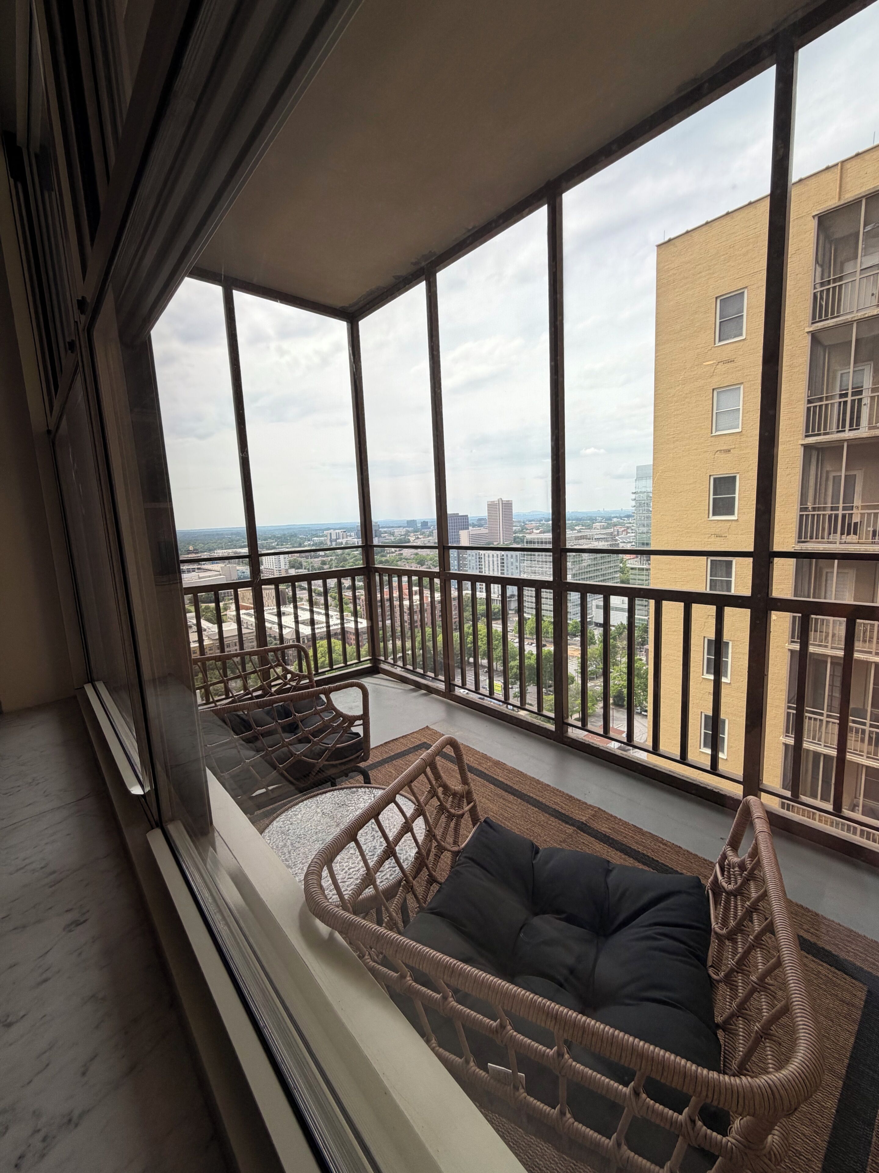 A balcony with a wicker sofa and a small table, overlooking a cityscape with buildings and a clear sky.