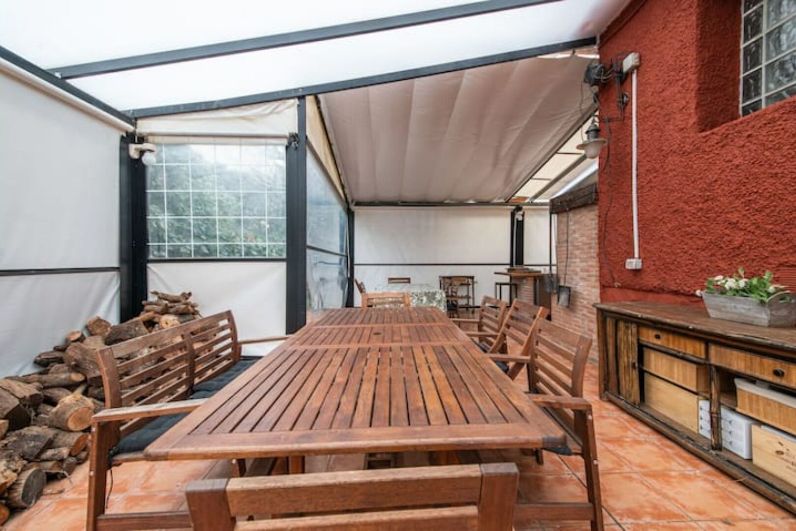 Un patio techado con una mesa y bancos de madera, un muro de ladrillo y una ventana con vista a árboles.