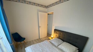 A bedroom with a bed, a chair, a bedside table with a lamp, and a door leading to another room.
