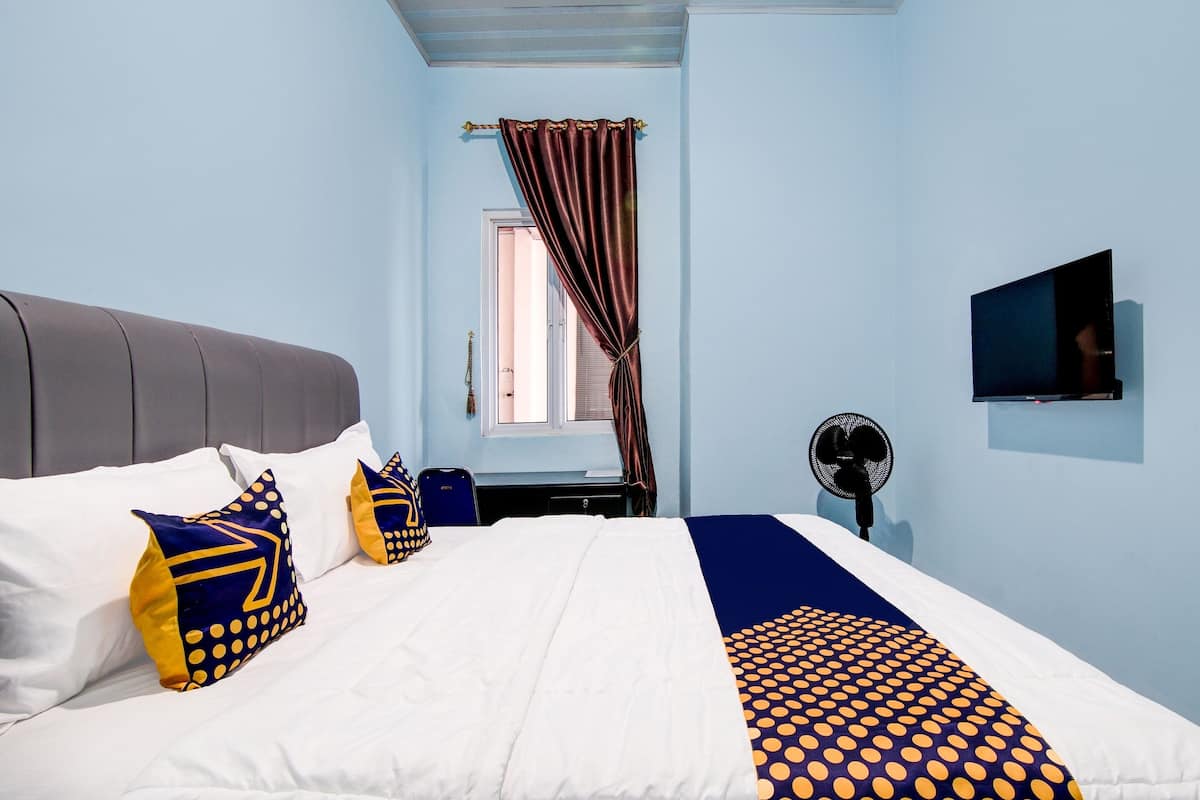 Een slaapkamer met een bed, kussens, een televisie, een ventilator en een raam met gordijnen.