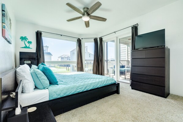 A bedroom with a bed, a TV, a ceiling fan, and a view of the outside.