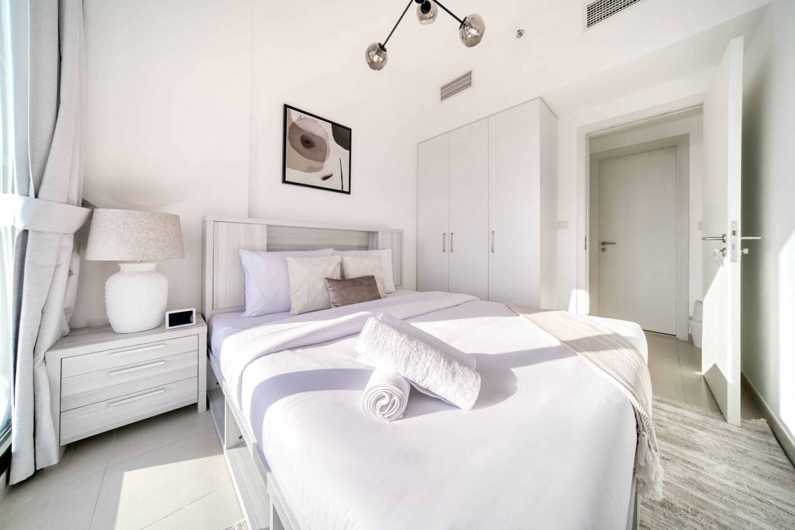 Ein modernes Schlafzimmer mit einem großen Bett, einem Nachttisch mit Lampe, einem Kleiderschrank und einem gerahmten Bild an der Wand.