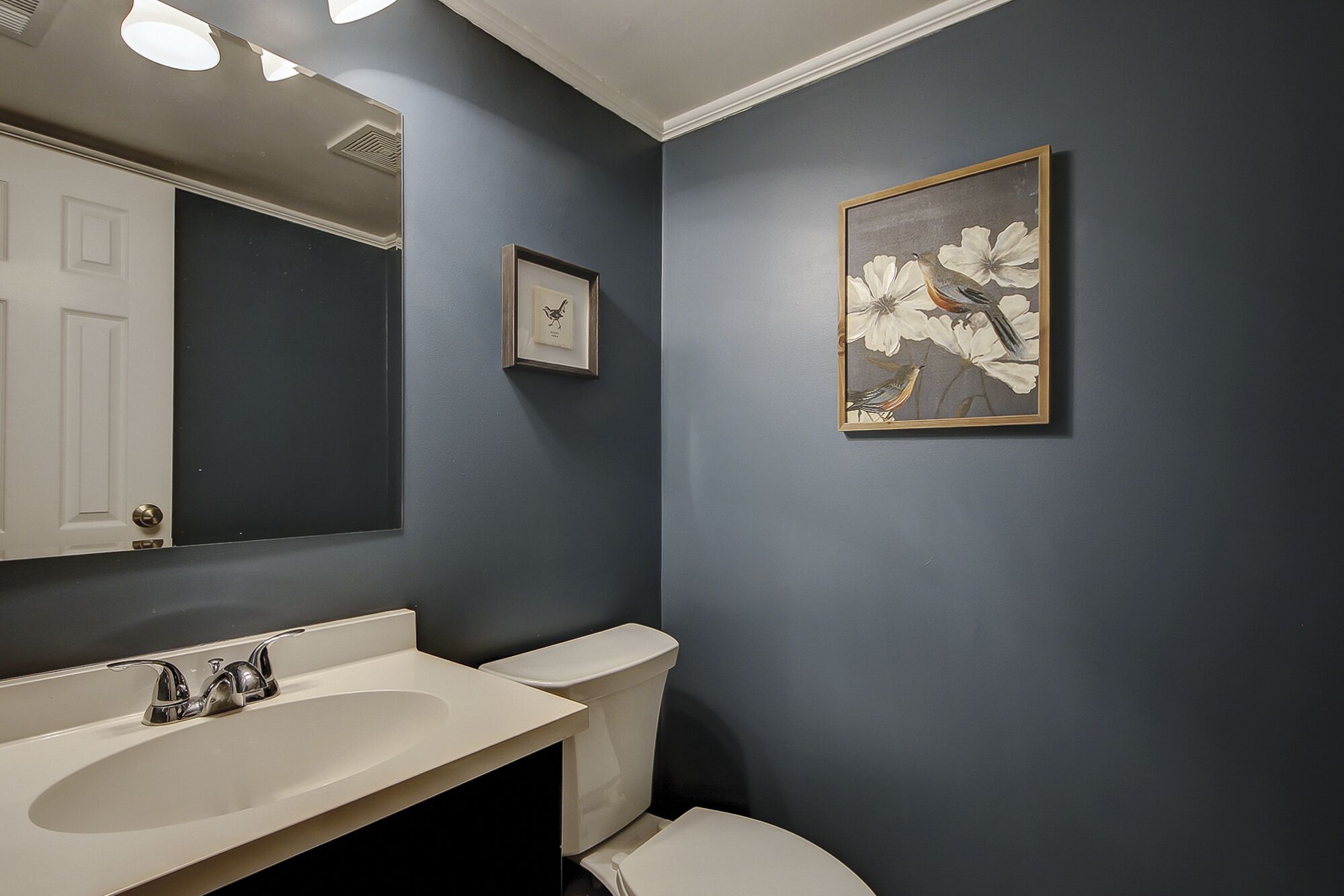 A bathroom with a dark blue wall, a white sink, and a toilet. There is a framed picture of birds on the wall.