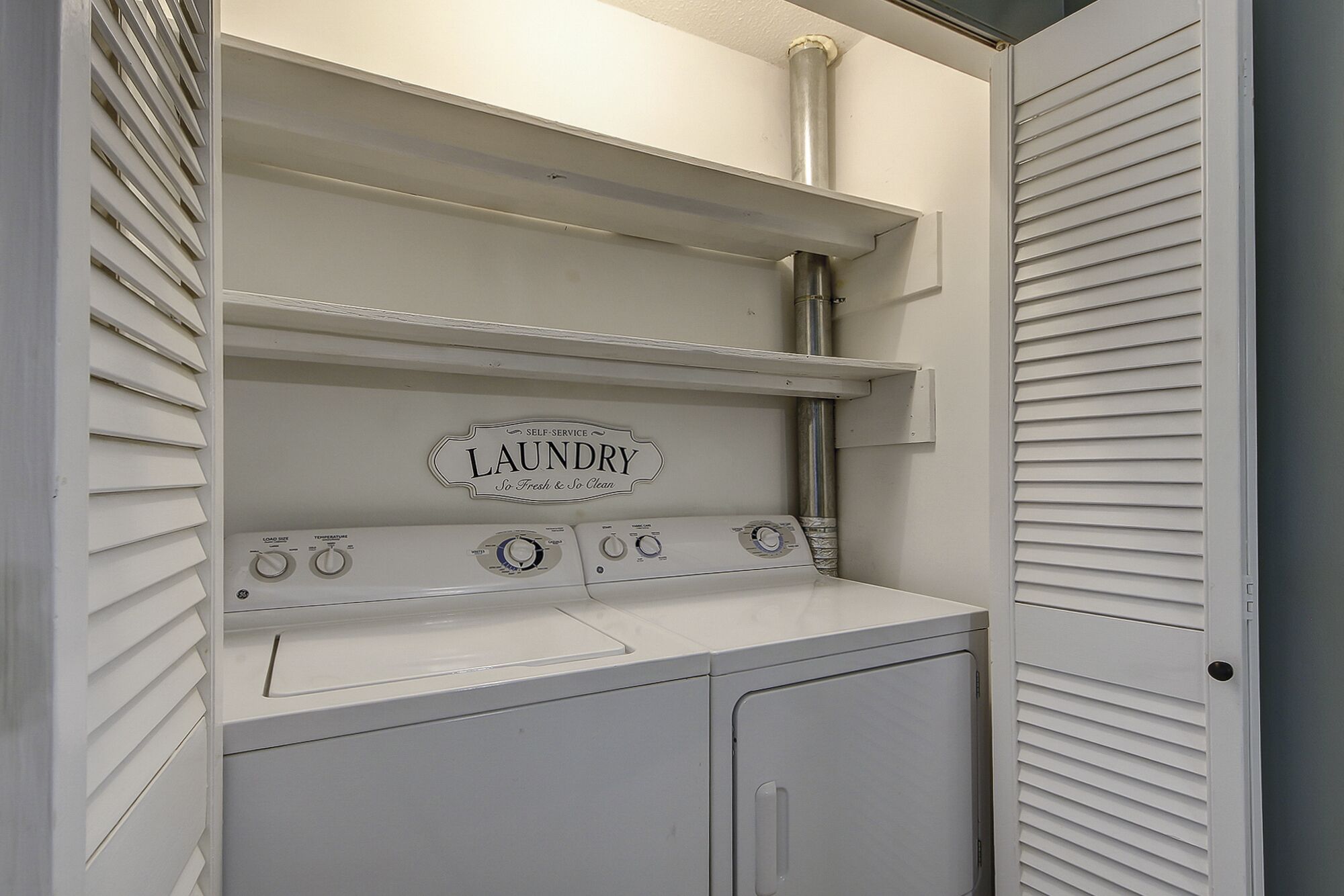 A laundry area with a self-service machine, a shelf above it, and a door with louvered panels.