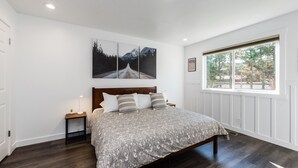Un dormitorio con una cama grande, una mesita de noche, una ventana con vista a árboles y un cuadro colgado en la pared.