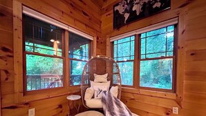 Une pièce chaleureuse aménagée dans un chalet, avec des murs en bois, de grandes fenêtres, un fauteuil en osier et un pouf rond.