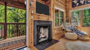 L’intérieur d’un chalet en bois, avec une cheminée, un téléviseur fixé au mur et une vue sur l’extérieur.