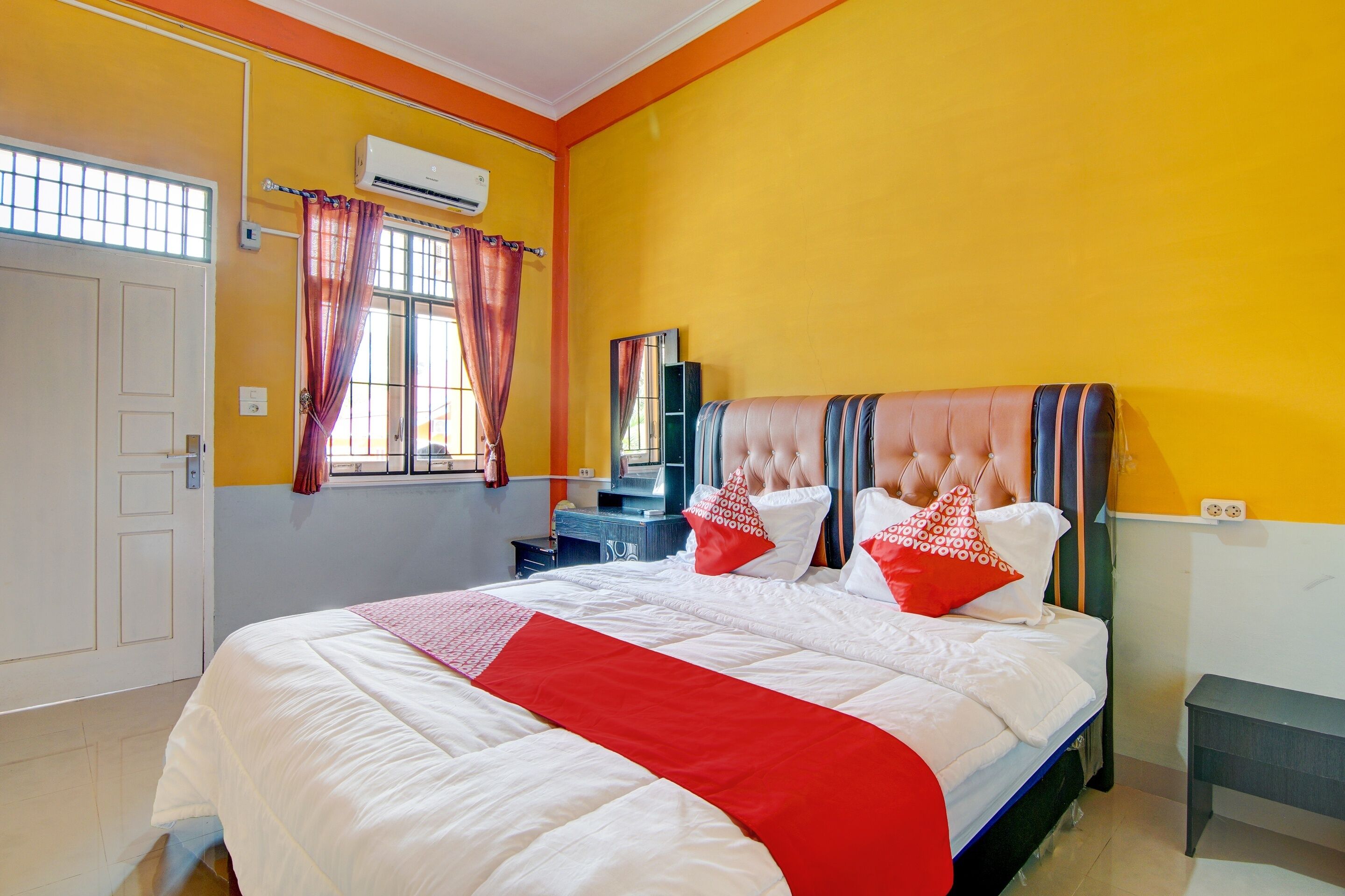 A hotel room with a bed, a vanity, an air conditioning unit, a window with curtains, and a door.