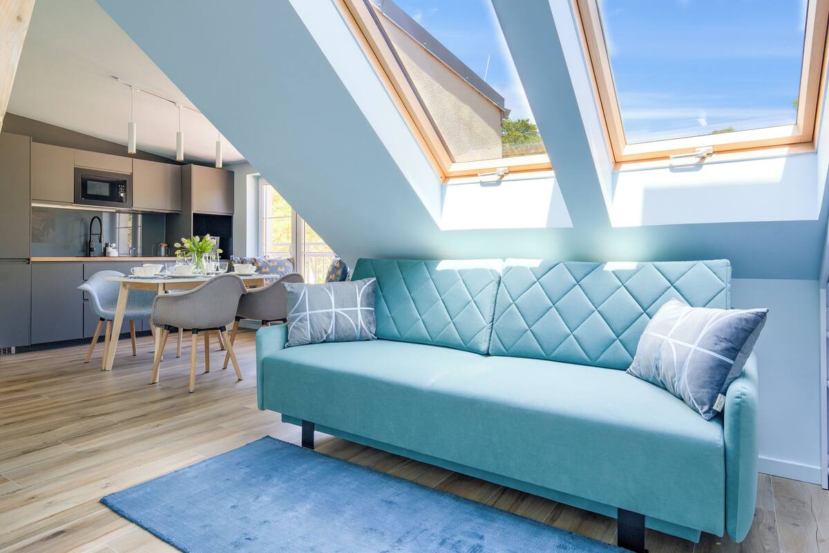 Un appartement moderne de style « loft » comprenant un coin repas, une cuisine et un espace de vie chaleureux avec un canapé turquoise et une lucarne.