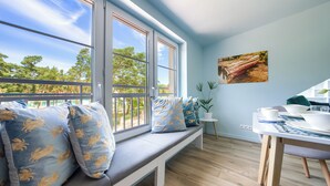 A room with a large window, a bench with patterned cushions, a dining table set with plates and cups, and a potted plant. - Apartamenty Sun & Snow Las i Morze (Trzebiatow)