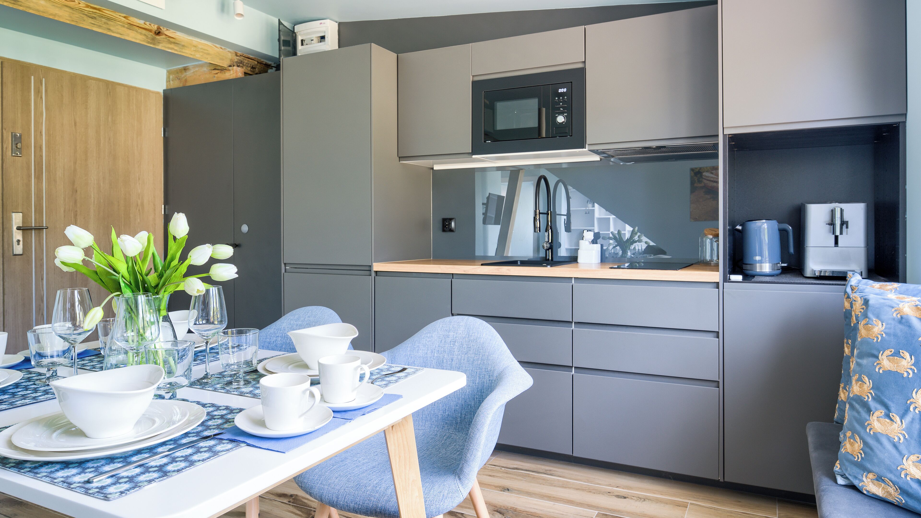 Une cuisine moderne avec des meubles gris, un four à micro-ondes et une machine à café. On y trouve une table à manger dressée avec de l’assiette blanche, des bols et des verres. Une chaise bleue est visible, et un vase avec des fleurs blanches est posé sur la table.