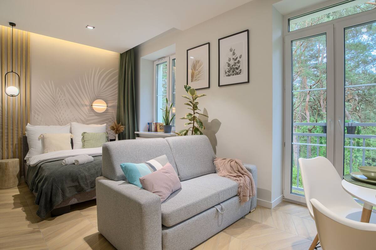 Une chambre moderne avec un lit, un canapé gris, une table à manger avec des chaises et de grandes fenêtres donnant sur la forêt.