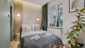 A modern bedroom with a bed, a bedside table, a radiator, a window with a view of trees, and a potted plant. - Apartamenty Sun & Snow Las i Morze (Trzebiatow)