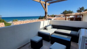 Un espace extérieur aménagé, abrité et doté d’une vue sur la mer, comprenant un canapé, des poufs et un ventilateur.