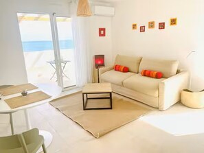 A modern living room with a sofa, a small table, a lamp, and a view of the sea through a glass door.
