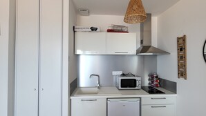 Une cuisine compacte avec des meubles blancs, un four à micro-ondes, un petit réfrigérateur, un évier et un luminaire suspendu en osier.