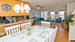 A spacious living area with a dining table set for four, a kitchen with a wooden island, and a balcony with a view. - Irish Mist, Oceanfront House in Emerald Isle, NC! (Emerald Isle)