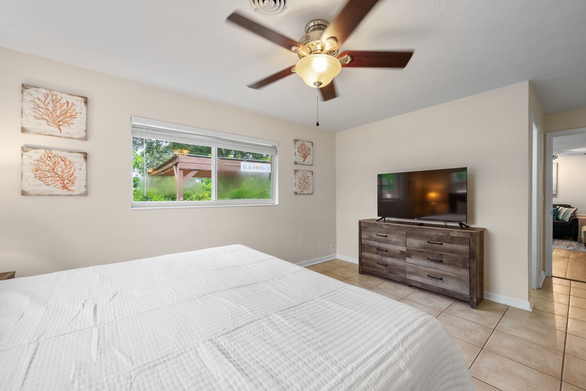 A bedroom with a bed, a TV, a ceiling fan, and a window with a view of a house.