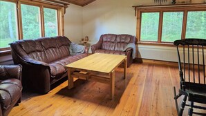 Een gezellige woonkamer met een bruine leren bank, een houten bijzettafeltje en een zwarte stoel. Grote ramen bieden uitzicht op het landschap.