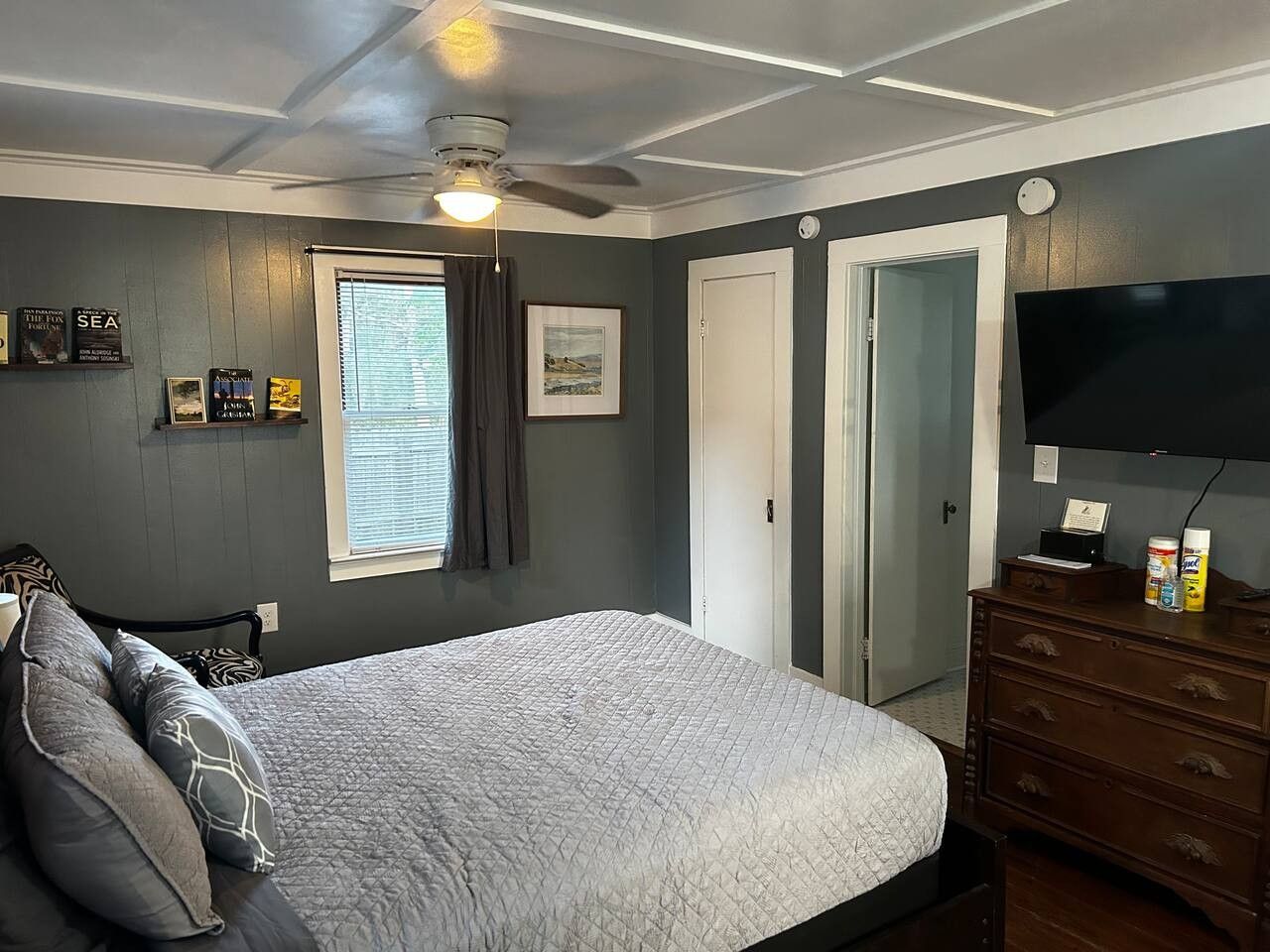 Quarto com cama, cômoda, ventilador de teto, janela com cortinas e televisão de tela plana.