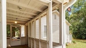 A covered porch with a wooden ceiling and white siding, leading to a door with a view of a garden. - Cute Cottage with Hot Tub-Historic District  (Ocala)