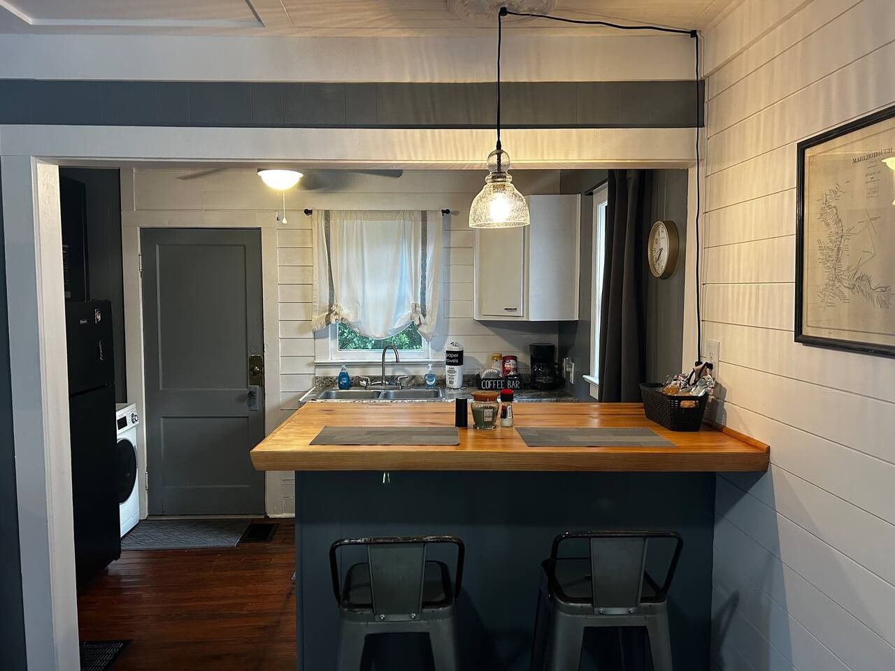 Cozinha compacta com ilha de madeira, dois banquetes, um espaço para café e um relógio na parede.