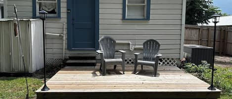 Une petite maison avec une porte et des fenêtres bleues, une terrasse avec deux chaises et un appareil de climatisation.