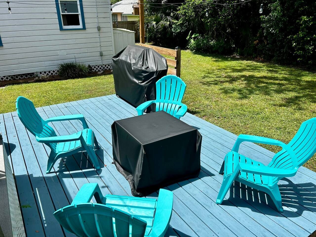 An outdoor deck with turquoise chairs and a covered table, surrounded by a grassy yard and a house.