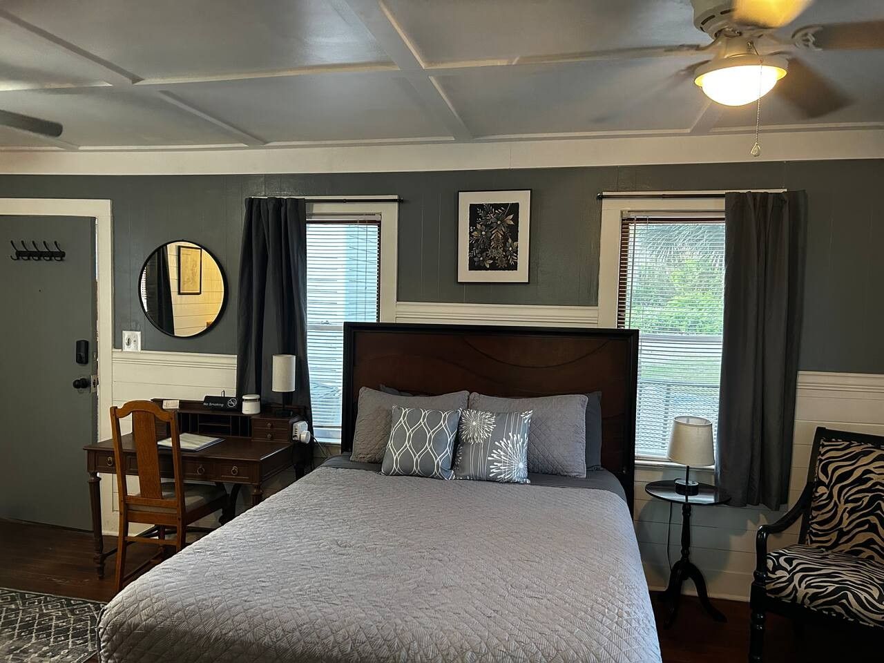 A bedroom with a large bed, a desk, a chair, a mirror, and two windows with curtains.