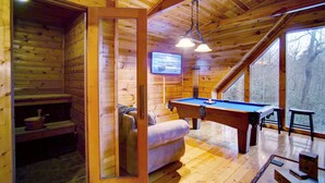 A wooden cabin interior with a pool table, a sofa, and a television.