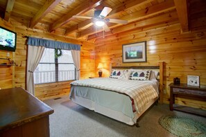 Quarto de cabana de madeira com cama, ventilador de teto, televisão e janela com cortinas.