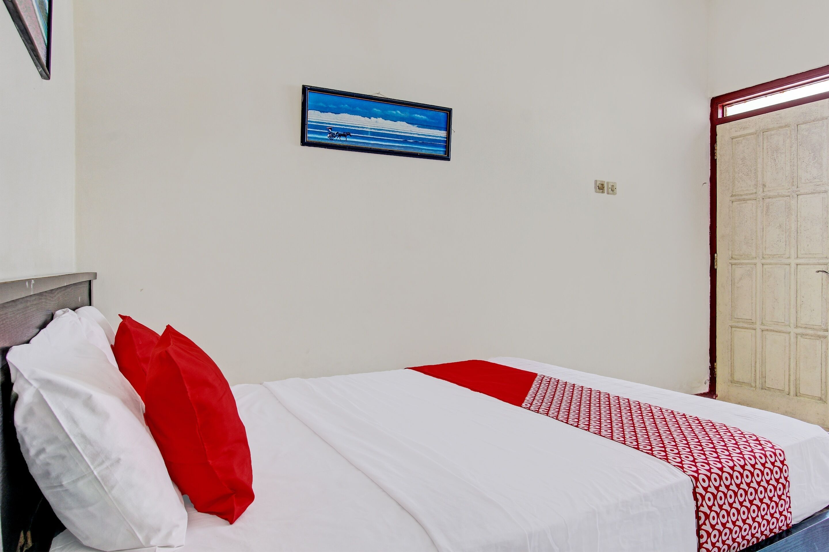 A bed with white and red bedding, a framed picture on the wall, and a door with a red frame.