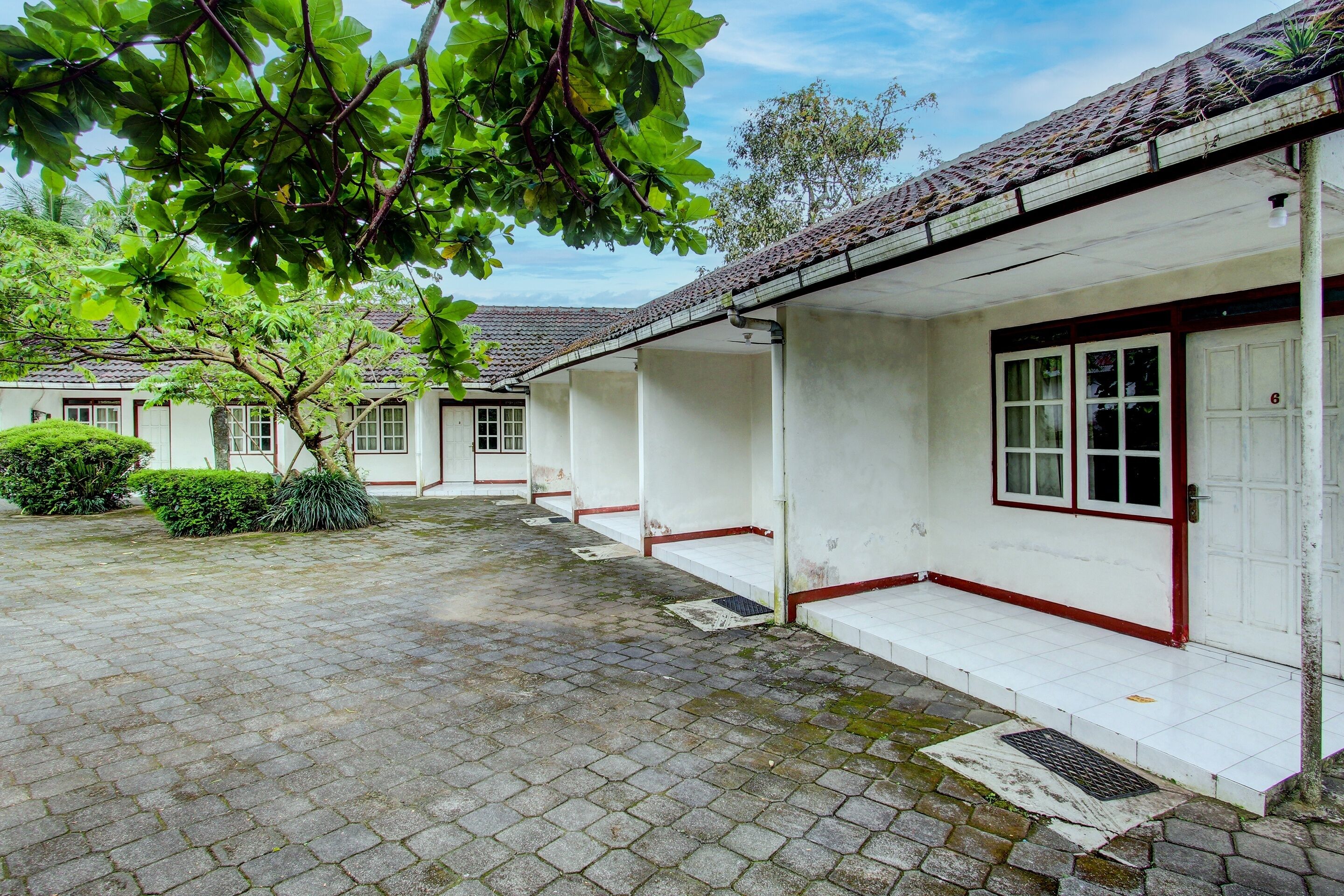 A courtyard with a paved walkway, white buildings with red trim, and a tree providing shade.
