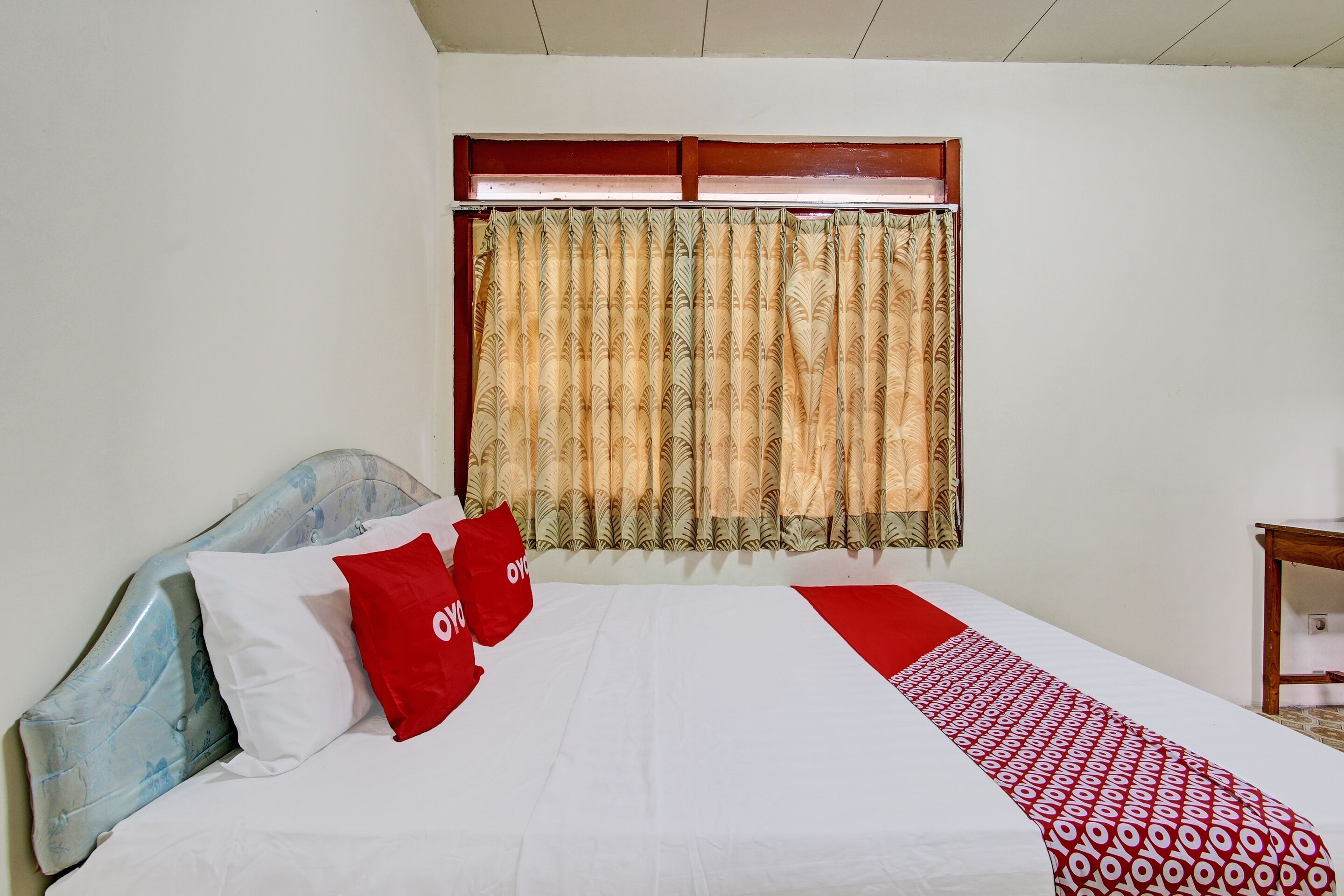 A hotel room with a bed, a headboard, a window with curtains, and a bedside table.