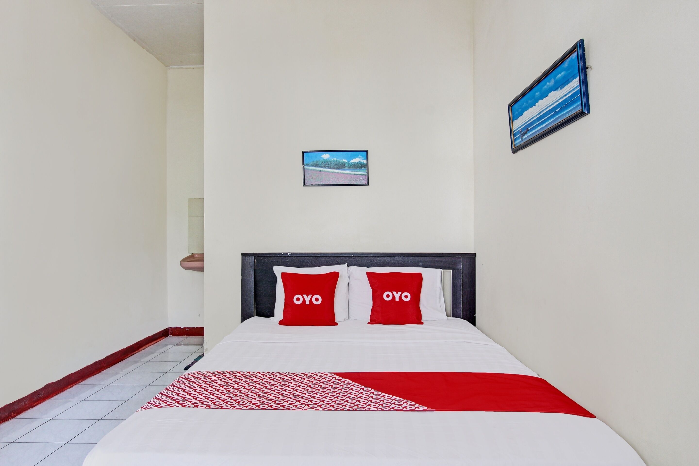 Une chambre d’hôtel avec un lit, deux oreillers rouges ornés du mot « OYO », une couvre-lit blanche et rouge, un petit tableau encadré accroché au mur et un sol carrelé.