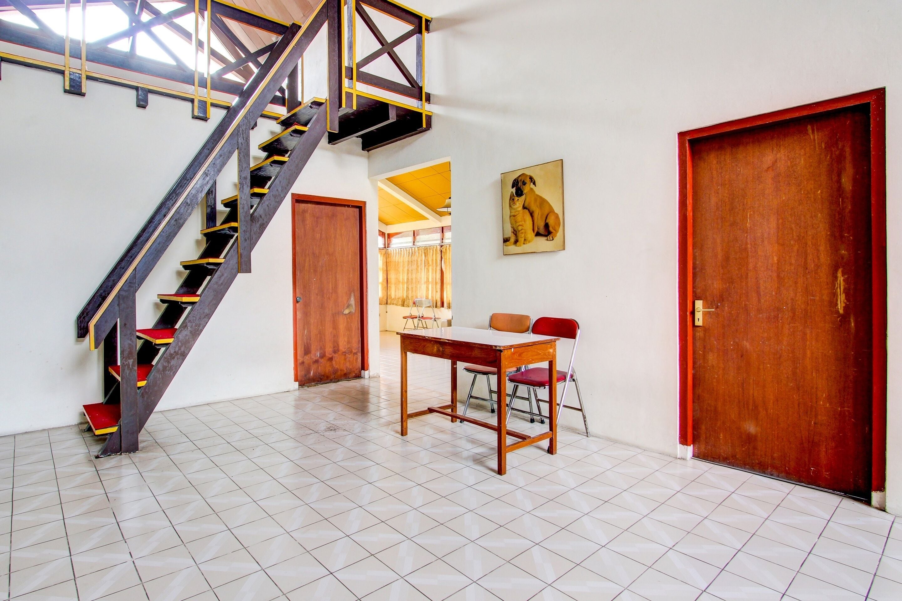 A staircase with a wooden handrail and red steps, a wooden table with two chairs, a wooden door, and a framed picture on the wall.