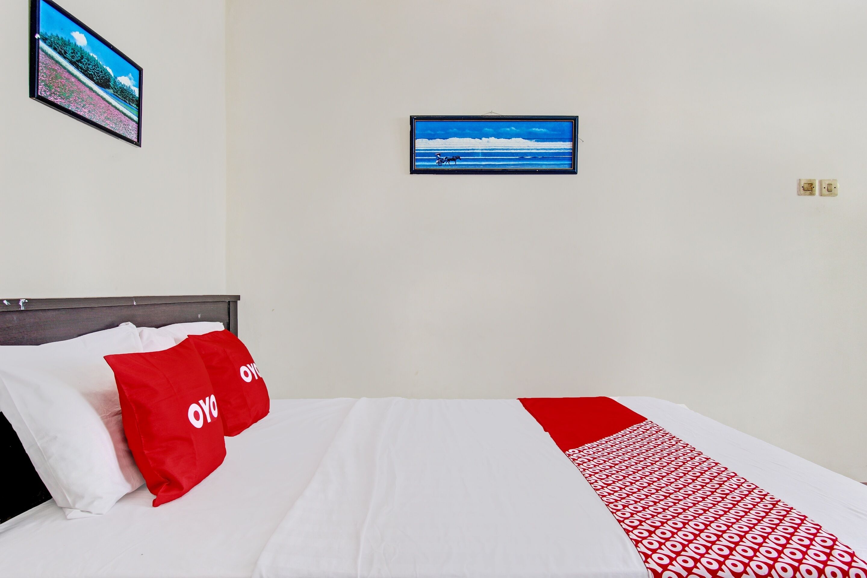 A hotel room with a bed, a red and white blanket, and two framed pictures on the wall.