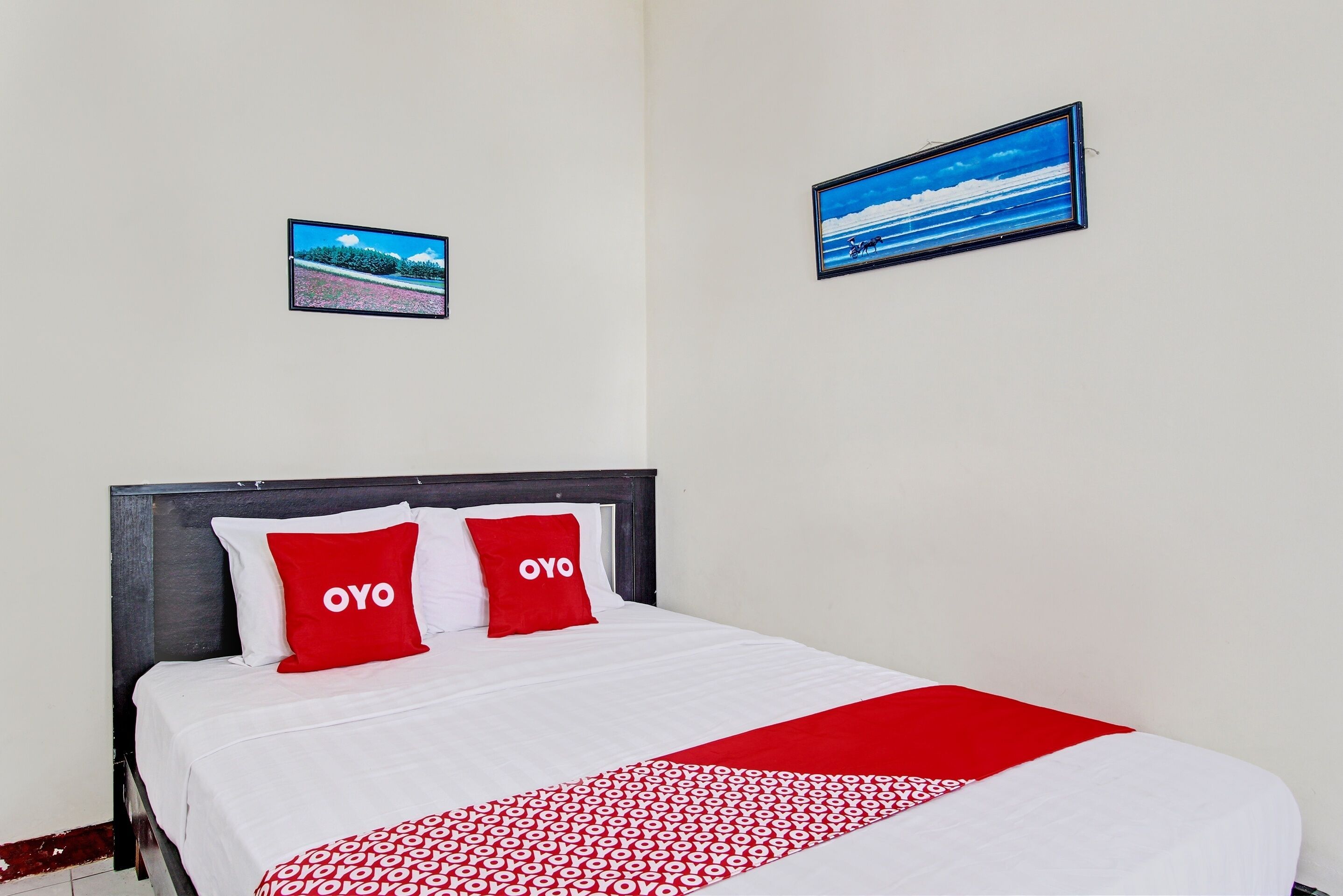 Et hotelværelse med en seng, to røde puder med teksten "OYO", et rødt og hvidt mønstret tæppe og to billeder i rammer på væggene.