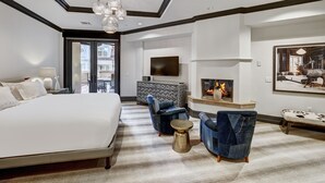 A modern hotel room with a large bed, a seating area with chairs and a coffee table, a fireplace, and a TV mounted on the wall.
