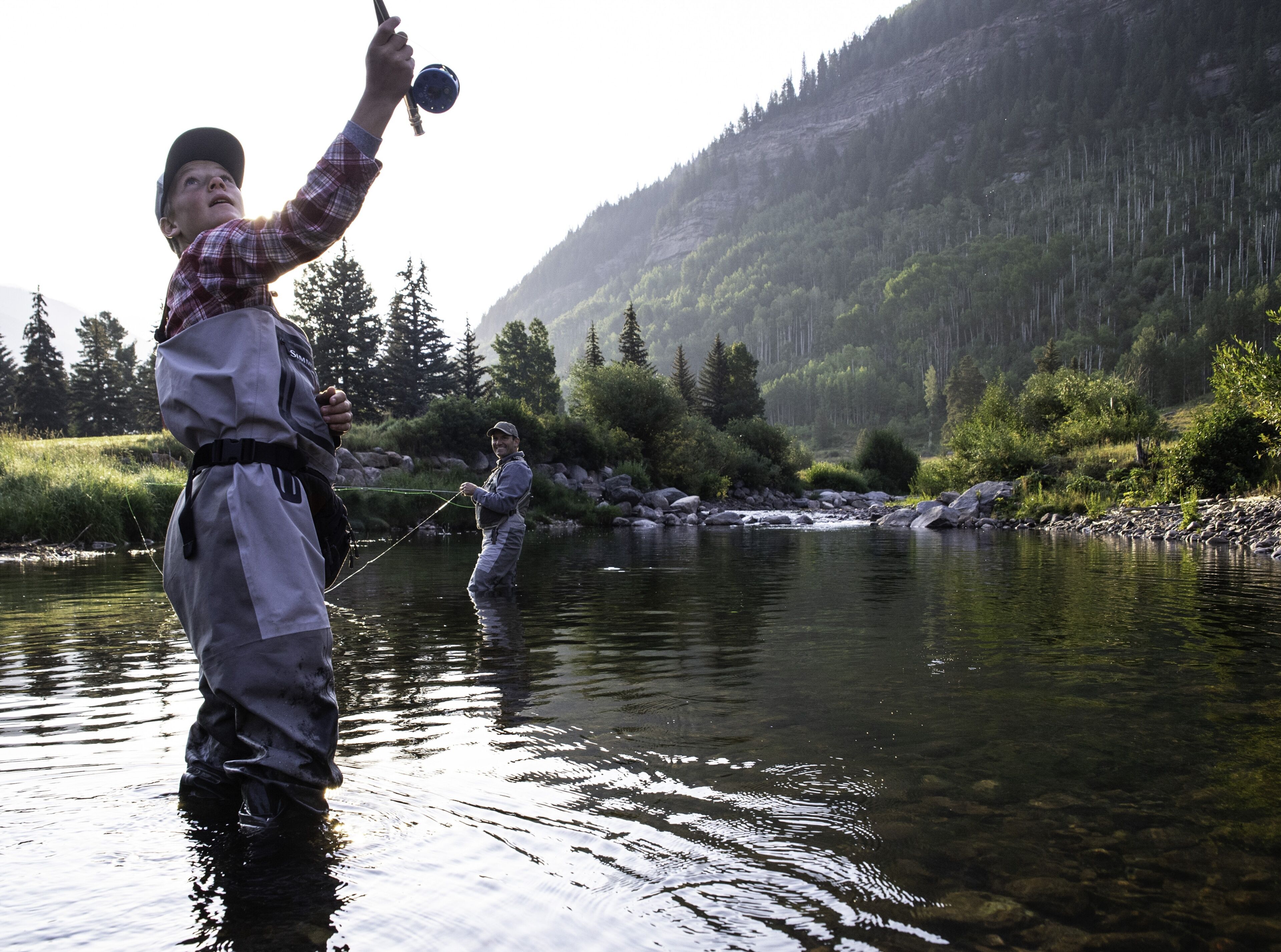 A person standing in a river with a fishing rod, another person in the background, and a mountainous landscape.