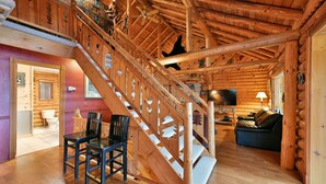 Interior de una cabaña de troncos con una escalera de madera, un comedor y una sala de estar con chimenea.