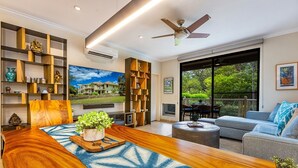A modern living room with a wooden coffee table, a large TV, and a balcony with outdoor furniture. - Grand Champions 29 1 Bedroom Condo (Kihei)